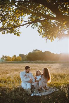 a family sitting on a blanket under a tree in the grass at sunset with their child