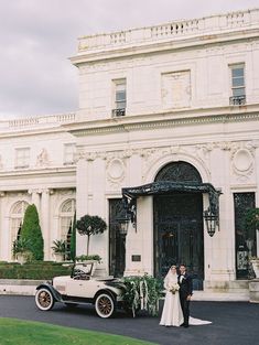 a bride and groom standing in front of an old fashioned car on the driveway of a mansion