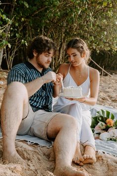 a man and woman sitting on the beach eating cake