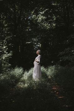 a woman standing in the middle of a forest wearing a white dress and looking up