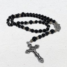 "A dramatic, bold, and rich Rosary, featuring jet black onyx for both the Hail Marys and Our Fathers. It also uses silver-plated nugget spacers to accent the beads, and an antique silver tone Miraculous Medal and Crucifix, made in Italy, as simple, elegant elements on the path of the Rosary. SPECIFICATIONS: * Hail Mary Beads: Smooth and Polished, Natural Black Onyx, 8 mm. * Our Father Beads: Smooth and Polished, Natural Black Onyx, 10 mm. * Centerpiece: Antique Silver Tone Miraculous Medal. * Crucifix: Antique Silver Tone Crucifix. Made in Italy. * Spacer and Accent Beads: Beautifully handmade Silver Plated Brass Nugget Chips. * Length: 17 1/4\" All Rosaries come in a handmade tapestry Rosary pouch. DESIGN STORY: Black Onyx has long been associated with new beginnings, healing, and regener St Michael Medal, Handmade Tapestries, The Rosary, Black Ink Tattoos, Miraculous Medal, Pure Beauty, Blue Agate, Simple Elegant, Agate Beads