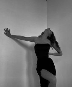 a woman in a black dress leaning against a wall with her hands on the wall