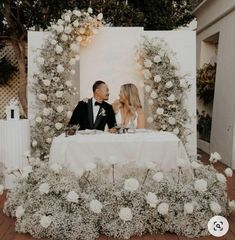 a man and woman sitting at a table in front of a white backdrop with flowers