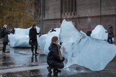 a woman kneeling down next to an ice sculpture on the sidewalk with people standing around it