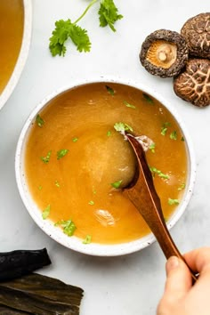 a person holding a spoon over a bowl of soup with mushrooms and parsley on the side