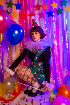 a woman dressed as a clown with balloons and confetti on her head sitting in front of a backdrop