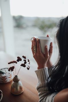 a woman sitting at a table holding a coffee cup in front of her face and looking out the window