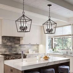 a kitchen island with stools and lights hanging from it's ceiling, in front of two windows