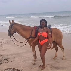 a woman in a bathing suit standing next to a horse on the beach near the ocean