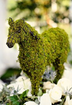 a moss covered horse standing on top of a white table cloth next to flowers and greenery