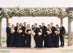 a large group of people in formal wear posing for a photo under an arch with flowers