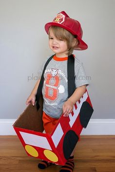 a young boy dressed up as a fireman holding a cardboard box with his hat on