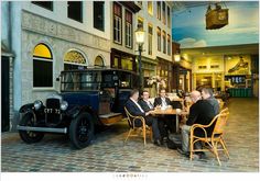 three men sitting at a table in front of an old car