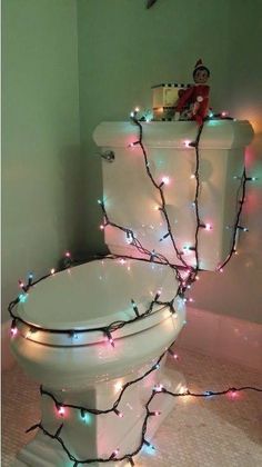 a white toilet covered in christmas lights next to a wall with a elf sitting on top of it