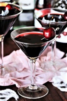 two martini glasses filled with dark colored liquid and topped with cherries on a wooden table