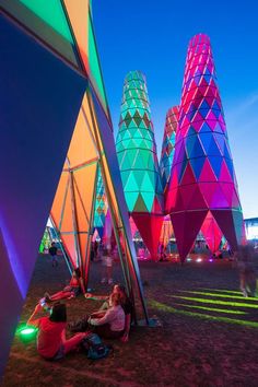 people are sitting on the ground in front of colorful structures at night, with bright lights shining