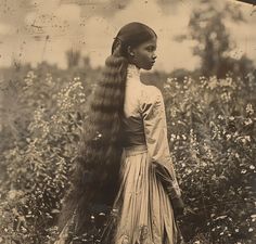 an old photo of a woman with long hair standing in a flowery field, looking to the side