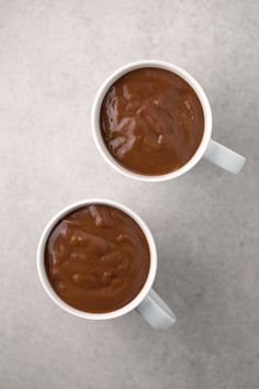 two white cups filled with chocolate sitting on top of a gray counter next to each other