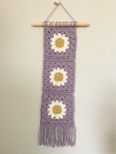 a purple crocheted scarf with white flowers hanging from a wooden hanger on a wall