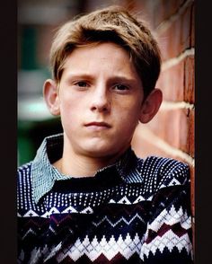 a boy leaning against a brick wall with his head turned to the side and looking at the camera