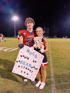 a man and woman holding a teddy bear in front of a sign on a football field