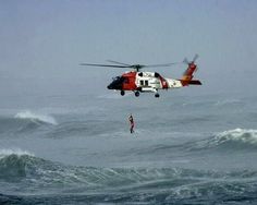 a red and white helicopter flying over the ocean next to a person standing on a surfboard