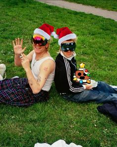 two people sitting on the grass wearing sunglasses and santa hats, one with his hand in the air