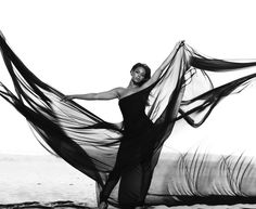 a woman is standing on the beach with her long black dress flowing in the wind