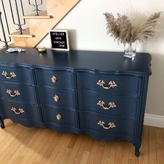 a blue dresser with gold leaf handles and drawers in front of a stair case next to a plant