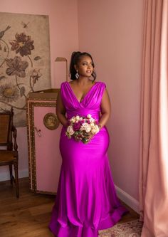 a woman in a purple dress standing next to a pink wall and holding a bouquet