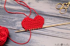 a red knitted heart sitting on top of a wooden table next to knitting needles