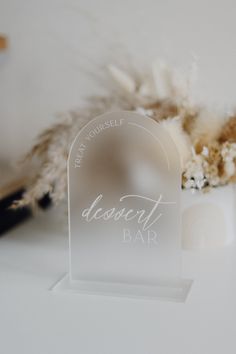 a clear acrylic sign sitting on top of a white table next to dried flowers