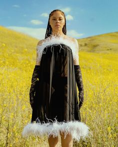 a woman standing in a field wearing a black dress with white feathers on it's shoulders