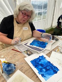 an older woman sitting at a table working on some art work with watercolors