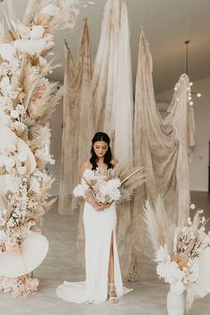 a woman in a white dress holding flowers and standing next to tall vases with feathers hanging from the ceiling