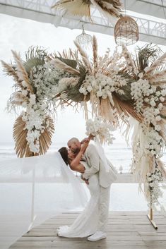 a bride and groom kissing under an arch decorated with white flowers, greenery and feathers