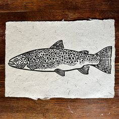 a drawing of a fish on a piece of paper with black and white ink, sitting on a wooden surface