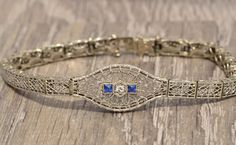 The Arvada Art Deco Filigree Bracelet with French Cut Sapphires and Diamond. The center is set with an old cut diamond with a single french cut sapphire on either side. Surrounding the stones is an intricate filigree pattern in the larger centerpiece. The bracelet has straight filigree links with an elaborate pattern of filigree with a central flower in each link. The bracelet has a push style clasp and is signed by the designer on the hidden safety. The bracelet is crafted in 14 karat white gol Vintage Hand Set Diamond Bracelet, Vintage Diamond Bracelet With Hand-set Diamonds, Classic Formal Bracelets With Intricate Design, Vintage Formal Diamond Bracelet With Hand Set, Vintage Diamond Bracelet With Hand Set For Formal Occasions, Vintage Hand Set Diamond Bracelet For Formal Occasions, Vintage White Gold Hand-set Bracelets, Art Deco Diamond Cut Bracelet For Wedding, Art Deco Diamond Bracelet As Gift