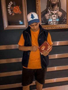 a young man in an orange shirt and blue hat is holding a book while standing next to two framed pictures