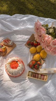 a table topped with cakes and flowers on top of a white sheet covered field next to fruit