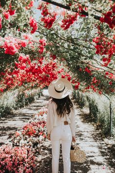 a woman standing in front of red flowers wearing a white suit and hat with her back to the camera