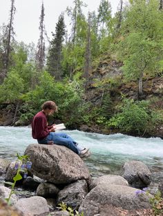 a young man sitting on top of a rock next to a river reading a book