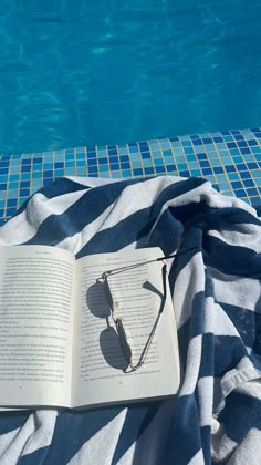 an open book sitting on top of a towel next to a swimming pool with sunglasses