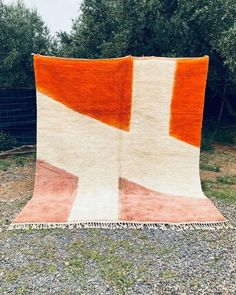 an orange and white rug sitting on top of gravel covered ground next to trees in the background