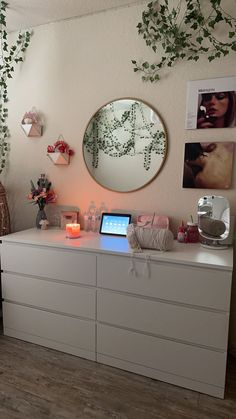 a white dresser topped with a laptop computer next to a mirror and plant hanging on the wall