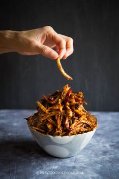 a hand holding a piece of food over a white bowl filled with shredded beef and chili