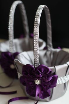 two silver baskets with purple flowers in them on a white tablecloth and ribbon tied around the handles