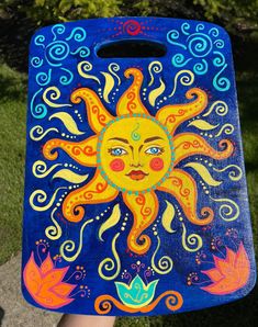 a painted sun on a blue board in front of some grass and trees, with one hand holding it