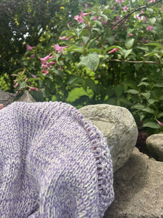 a purple and white knitted blanket sitting on top of a rock next to flowers