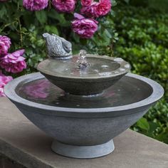 a birdbath sitting on top of a water fountain surrounded by pink flowers and greenery